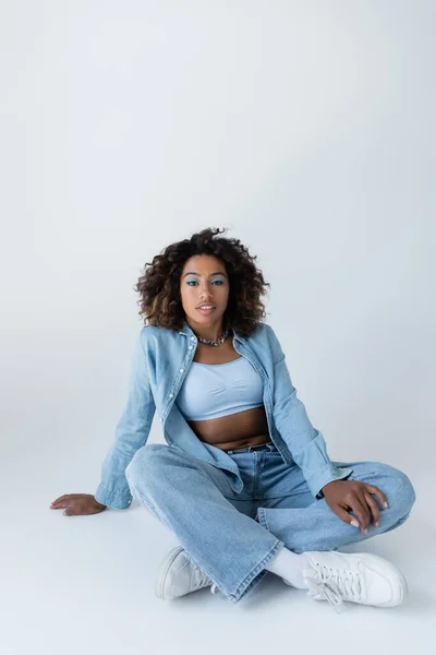 Brunette african american woman in blue denim clothes looking at camera while sitting on grey background — стокове фото