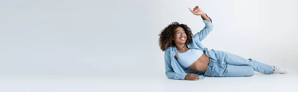 Щаслива афроамериканська жінка в джинсовому одязі, що лежить і вказує пальцем на сірому фоні, банер — стокове фото