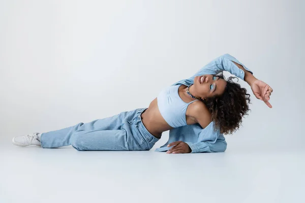 Повна довжина чуттєвої афроамериканської жінки у верхній частині білого врожаю і джинси, що лежать із закритими очима на сірому фоні — стокове фото