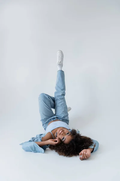 Smiling african american woman lying on back with raised legs on grey background — Fotografia de Stock