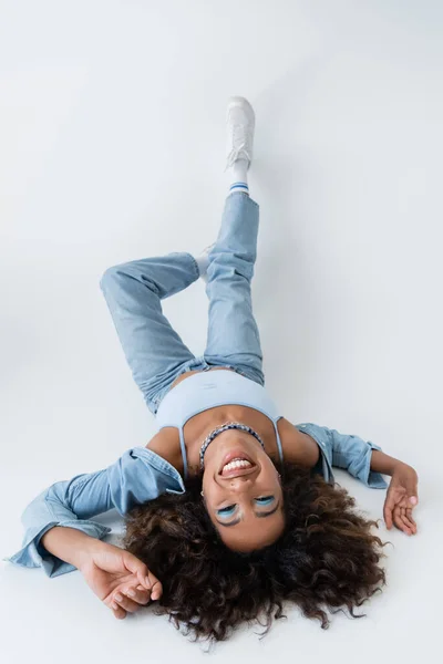 Високий кут зору щасливої афроамериканки в джинсовому одязі, що лежить на сірому фоні — стокове фото