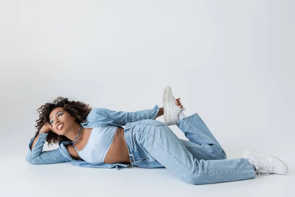 African american woman in trendy denim clothing looking away while lying on grey background — Stock Photo