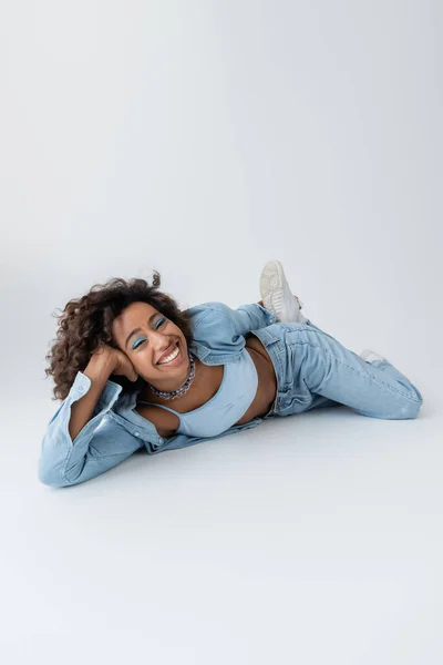Збуджена і стильна афроамериканка в джинсовому одязі, лежить на сірому фоні — стокове фото