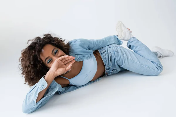 Cheerful african american woman in blue jeans covering mouth with hand while lying on grey background — Fotografia de Stock