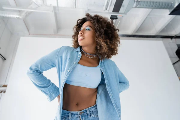 Trendy african american woman in white crop top and denim shirt looking away on white background - foto de stock