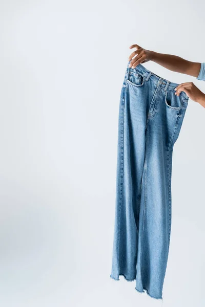 Partial view of african american woman holding blue and stylish jeans isolated on grey - foto de stock