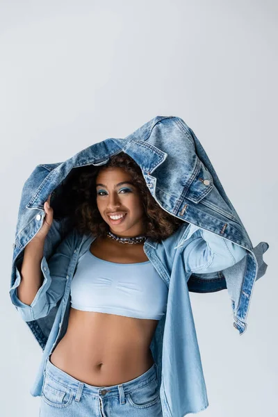 Smiling african american woman in white crop top posing with denim jacket isolated on grey — Photo de stock