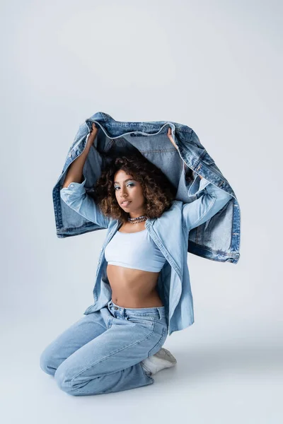 African american woman with denim jacket sitting on haunches on grey background — стокове фото