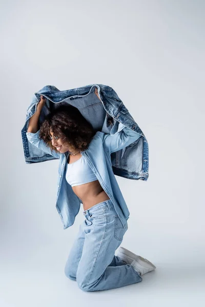 Happy african american woman posing with denim jacket while standing on knees on grey background — Stockfoto