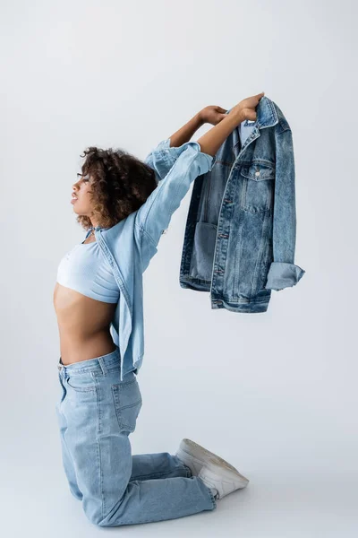 Side view of african american woman holding denim jacket while standing on knees on grey background — Stockfoto