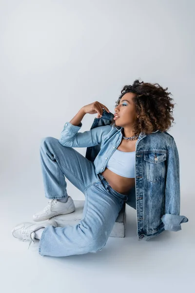 Full length of stylish african american woman in white sneakers and blue denim clothes sitting on grey background — Stock Photo
