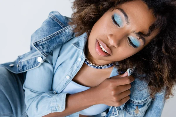 Sensual african american woman with blue eye shadows posing with denim garments isolated on grey - foto de stock