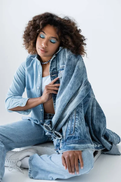 Trendy african american woman with makeup and closed eyes sitting in denim clothes on grey background — Foto stock