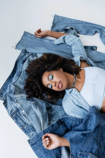 Top view of african american woman in beads lying near denim clothes on grey background — стокове фото