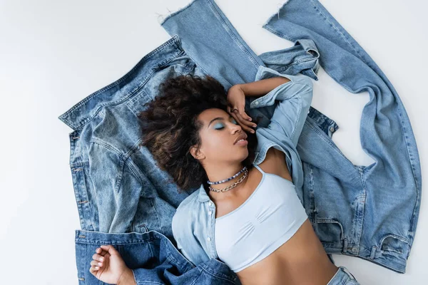 Top view of brunette african american woman lying near denim jacket and jeans on grey background — Stockfoto
