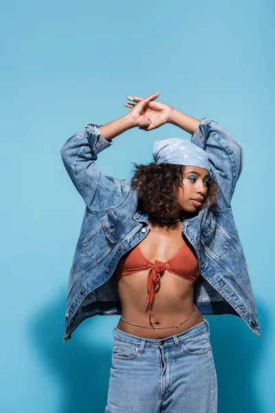 Fashionable african american woman in bandana and denim jacket standing with hands above head on blue background — Stockfoto