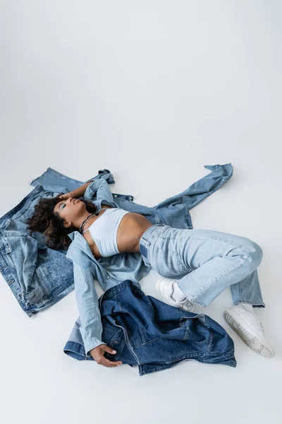High angle view of african american woman lying near denim garments on grey background - foto de stock