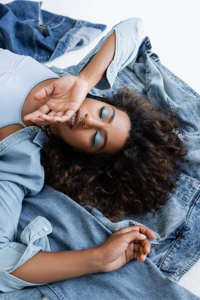 Зверху на афроамериканську жінку, що лежала з рукою біля обличчя біля денімського одягу на сірому фоні. — стокове фото
