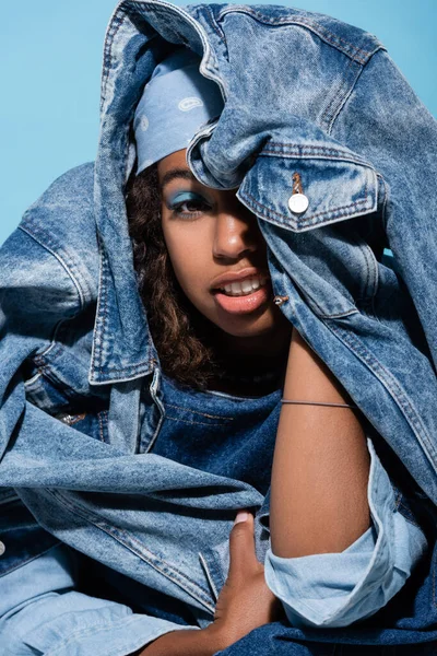 African american woman with makeup obscuring face with denim jacket isolated on blue — Stockfoto