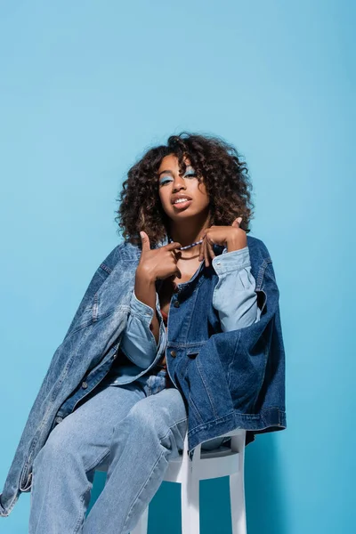 African american woman in denim clothes touching necklace while sitting on blue background — Photo de stock