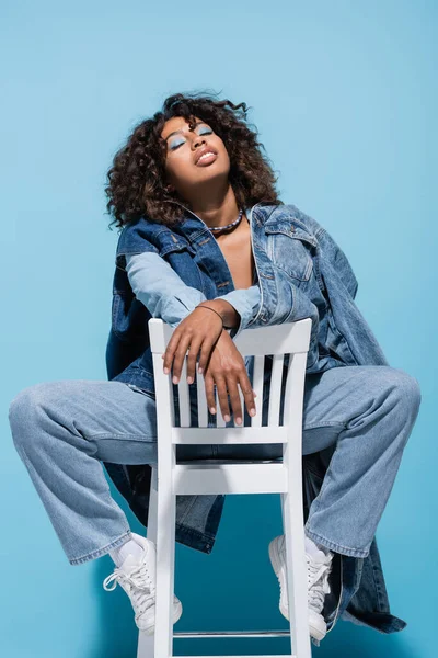 Trendy african american woman in denim clothes posing on chair with closed eyes on blue background — Fotografia de Stock