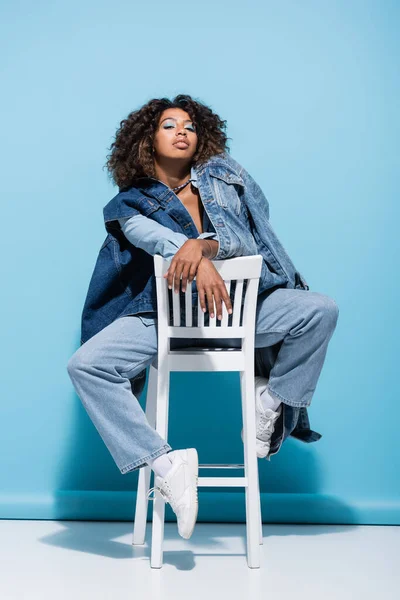 На синьому фоні жінка - афроамериканка, яка кладе на стілець денімний одяг. — стокове фото