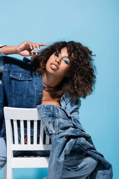 Trendy african american woman in denim clothing looking at camera on chair isolated on blue — стоковое фото