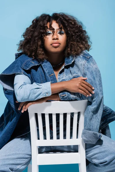 Stylish african american woman with wavy hair sitting on chair with crossed arms isolated on blue - foto de stock