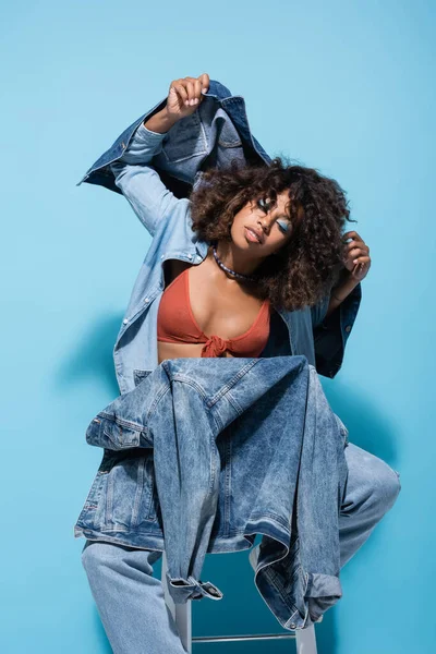 Модна афроамериканська жінка позує з джинсовим одягом на синьому фоні — стокове фото