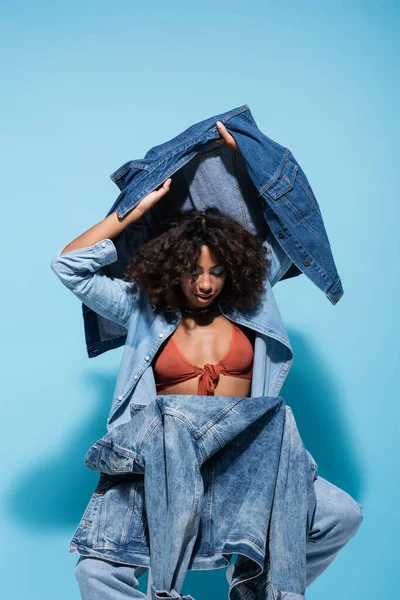 Trendy african american woman holding denim jacket and vest while sitting on blue background — Foto stock