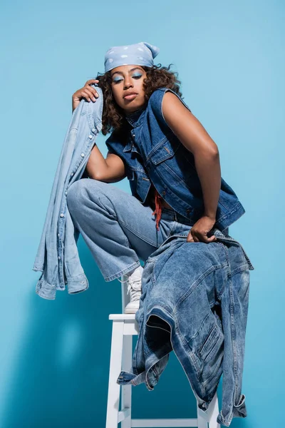 African american woman posing on chair with denim garments on blue background — Stock Photo