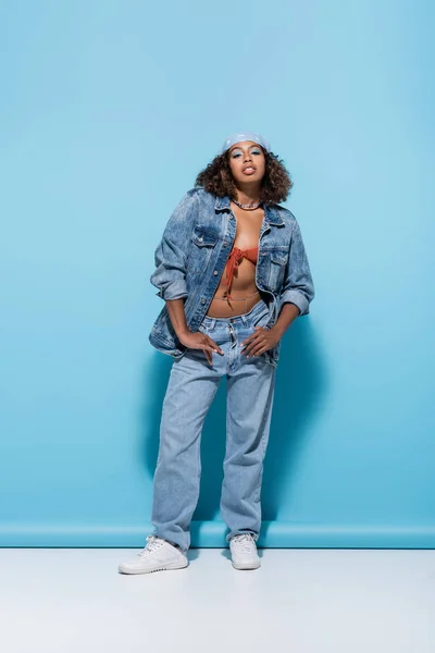 Full length of african american woman in denim clothes posing with thumbs in pockets on blue background — стокове фото