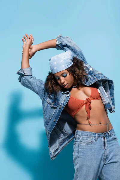 African american woman in bandana and trendy denim clothes posing with raised hands on blue background — Stockfoto