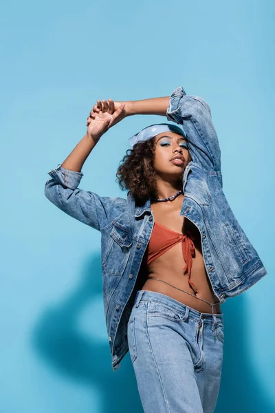Fashionable african american woman in denim jacket and bandana standing with hands above head on blue background — Stockfoto