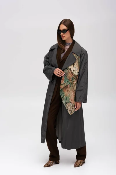 Full length of brunette teenage model in stylish sunglasses and trendy trench coat on grey — Stockfoto