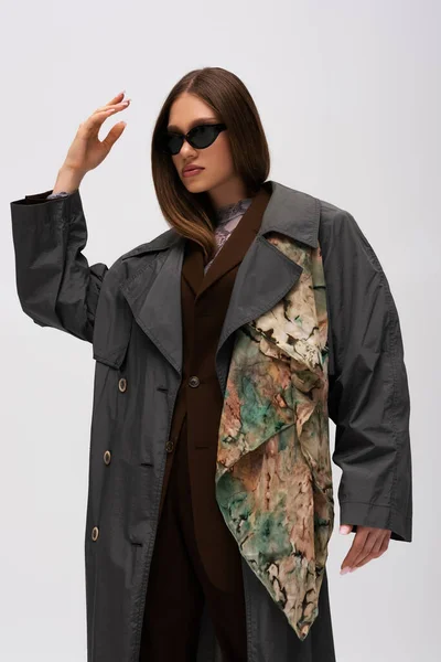 Стильна модель підлітка в сонцезахисних окулярах і модна траншея пальто позує ізольовано на сірому — стокове фото
