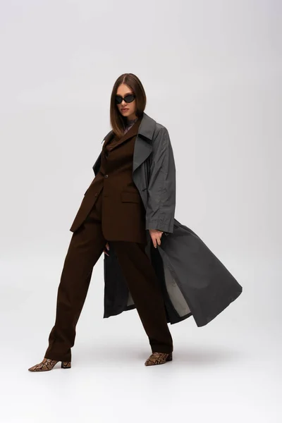 Full length of stylish teenage model in sunglasses and trendy trench coat walking on grey — Fotografia de Stock