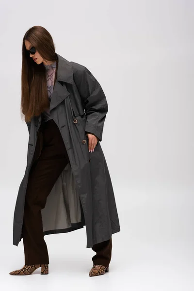 Full length of teenage model in sunglasses and trench coat posing on gray — Fotografia de Stock