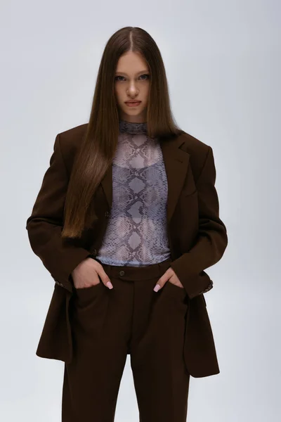 Гарна модель підлітка в коричневому високоякісному костюмі позує руками в кишенях ізольовані на сірому — стокове фото
