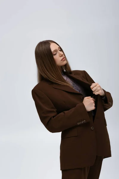 Pretty teenage model adjusting brown high-quality blazer while posing isolated on grey — Photo de stock