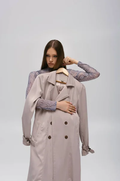 Pretty teenage model holding recyclable trench coat on hanger isolated on grey — Stockfoto