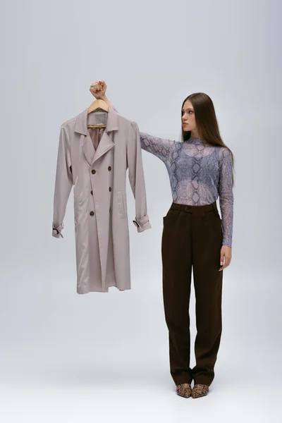 Full length of teenage model looking at recyclable trench coat on hanger on grey — Stockfoto