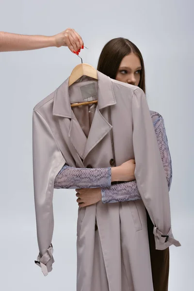 Модель-подросток, обнимающая стильное пальто, в то время как женщина, держащая вешалку, изолирована на сером — стоковое фото
