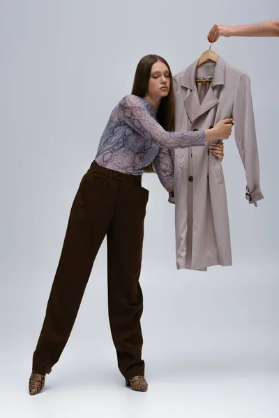 Full length of teenage model hugging stylish trench coat while woman holding hanger on grey — Stock Photo