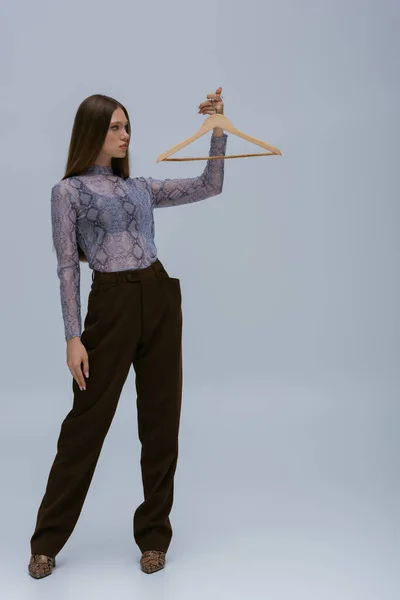 Full length of brunette teenage model posing with wooden hanger on grey — Fotografia de Stock