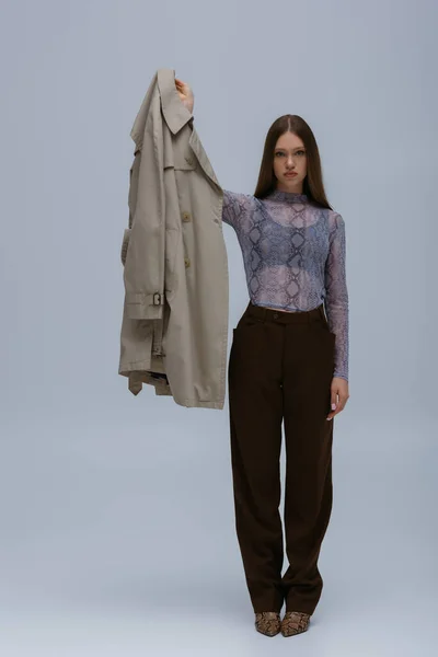 Full length of teenage model posing with outstretched hand and holding stylish trench coat on grey — Photo de stock