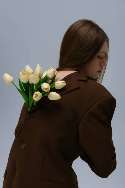 Вид сбоку модели-подростка в коричневом блейзере с тюльпанами, позирующими на сером — стоковое фото