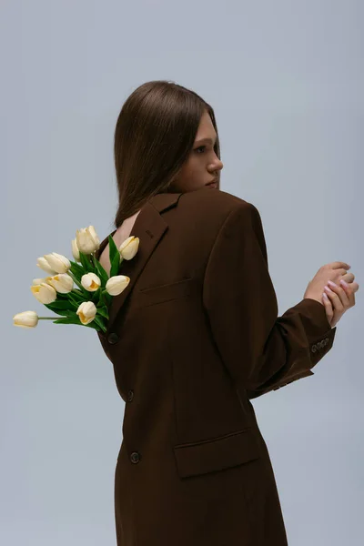 Вид сбоку симпатичной модели в коричневом блейзере с тюльпанами, позирующими изолированными на сером — стоковое фото