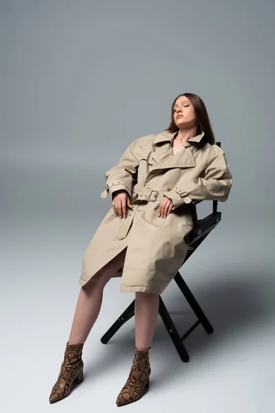 Повна довжина моделі з закритими очима в стильному траншеї пальто позує на стілець сірого кольору — стокове фото