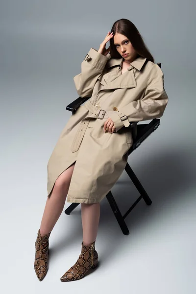 Full length of model in stylish trench coat posing on chair of grey — Stockfoto